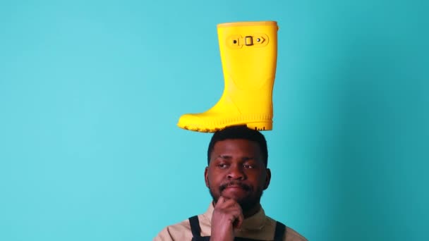 Nigerijský zahradník muž ukazuje žlutou gumovou obuv v modrém studiu připraven na podzimní jarní sezónu — Stock video