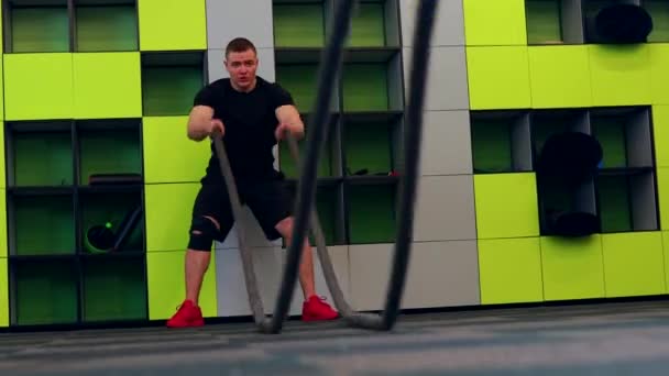 Hombre atleta masculino joven y guapo haciendo ejercicios para los músculos del cuerpo con cuerdas de batalla en gimnasio de fitness con fondo amarillo — Vídeos de Stock