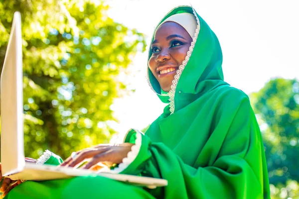 Junge schöne muslimische Geschäftsfrau mit modernem Make-up trägt grünen Hijab und benutzt Laptop, während sie im Sommerpark sitzt — Stockfoto