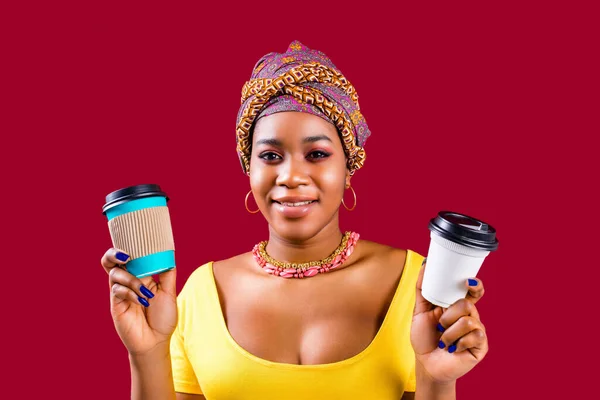 Змішана жінка в жовтій футболці та етнічному турбані над головою, показуючи паперовий стаканчик з африканською кавою, щоб вийти назовні на червоному студійному фоні — стокове фото