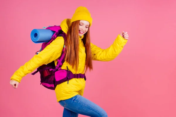 Female hiker with a backpack looking at the camera and smiling isolated on pink studio background.she wearing yellow windbreaker waterproof jacket and knitted hat