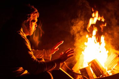 Yaz gecesi kamp yaparken ateşin yanında oturan genç adam.