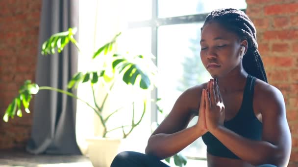 Mulher africana americana em sutiã desportivo preto está sentado postura de lótus no chão tapete de ioga olhos fechados e ouvir um áudio meditar mantra mãos namaste — Vídeo de Stock
