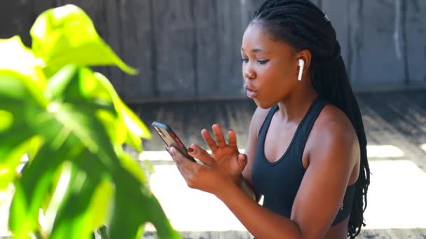 Латиноамериканка с афро-косичками слушает музыку от беспроводных ушных капсул носить спортивную одежду черный бюстгальтер и леггинсы на солнце в помещении — стоковое видео