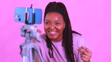 Koyu tenli kadın gülüyor ve pembe duvar arkasında sosyal medya hikayeleri çekiyor.