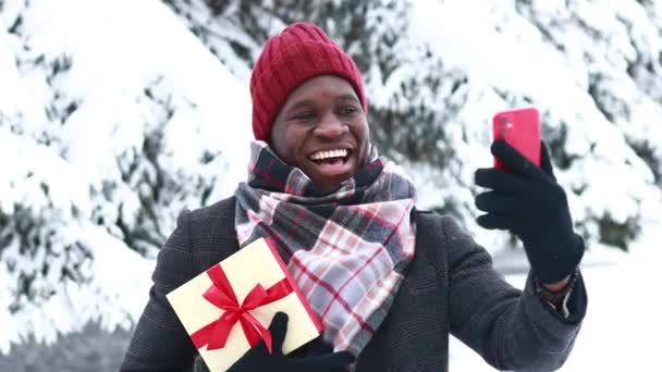 Polyetnische vrolijke man in stijlvolle jas en hoed met geschenkdoos met rood lint in winterpark, valentijnsdag concept. Hij praat met familie via videogesprek. — Stockvideo
