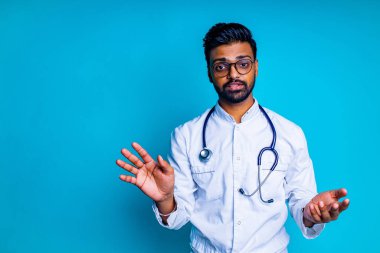 Hintli sakallı kardiyolog, mavi stüdyo arka planında stetoskobu olan, beyaz tıbbi önlüklü, gözlüklü adam.