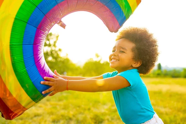 Positive multikulturelle Rennen kleines Mädchen mit afro lockigem Haar hält Regenbogenballon im Sommerpark — Stockfoto