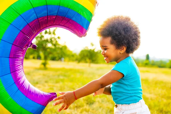 Süße afrikanisch-amerikanische kleines Mädchen mit lockigem Afro-Haar spielt Regenbogenballon im Freien — Stockfoto