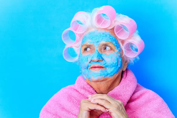 Gammal kvinna wirh rosa papiljotter på huvudet och blå hyaluron skönhet mask på ansiktet bära bomull mantel, förbereda sig för en dag Alla hjärtans dag i studio bakgrunds — Stockfoto
