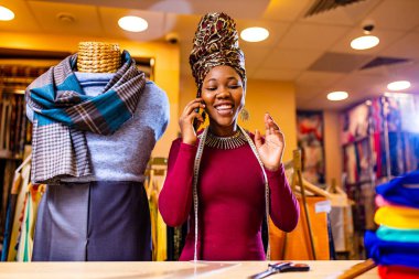 tanzanian woman with snake print turban over hear working in fabrics shop calling to client by smartphone clipart