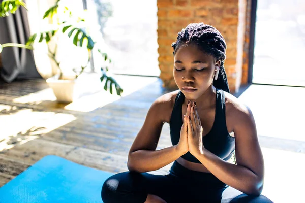 African american woman in black sporty bra is sitting lotus pose on yoga mat floor closed eyes and listening an audio meditate mantra hands namaste — Stock Photo, Image