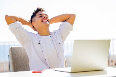 Kumsaldaki kafede dizüstü bilgisayarla çalışan beyaz tişörtlü genç bir adam.