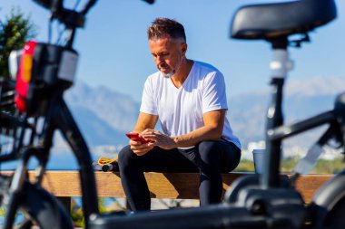 Beyaz tişörtlü yakışıklı adam, tepede güzel manzaralı eko scooterın yanında oturuyor.