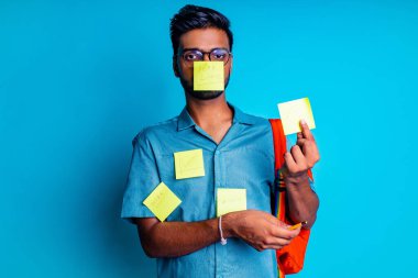 Stüdyo arka planında turuncu gökkuşağı sırt çantalı mavi pamuklu gözlüklü yakışıklı, ayı gözlü Hintli adam.