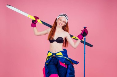 Çekici gülümseyen kızıl saçlı, güneş gözlüklü, bikinili ve çiçekli kızıl saçlı kayakçı fotokopi çekilen pembe arka planda kayakla poz veriyor.