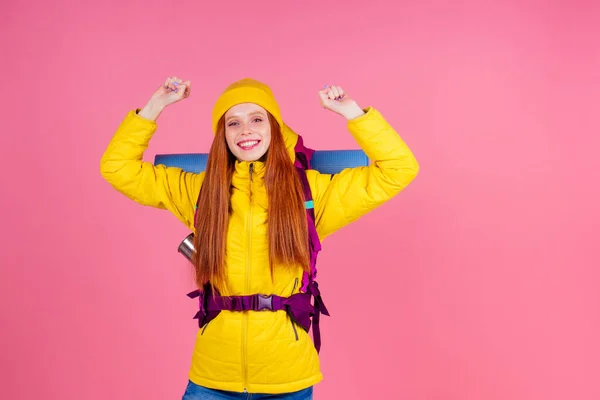 Female hiker with a backpack looking at the camera and smiling isolated on pink studio background.she wearing yellow windbreaker waterproof jacket and knitted hat