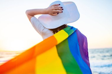 Saman beyaz şapkalı kadın elinde neşeli bir gökkuşağı bayrağı, kumsalda rüzgarda dalgalanıyor altın gün batımıyla. .