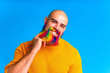 Renkli sakallı havalı adam kameraya bakıyor ve stüdyonun mavi arka planında harika bir ruh hali içinde..