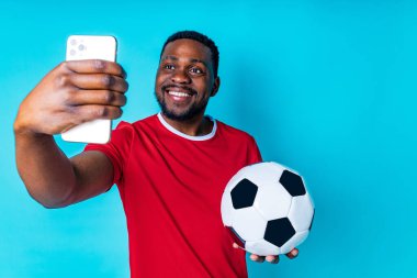 Stüdyonun mavi arka planında telefonu ve futbol topu olan Latin bir adam..