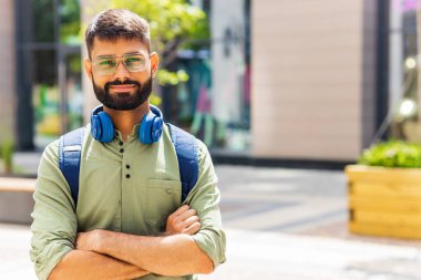 Mavi kulaklıklı ve sırt çantalı Hintli öğrenci güneşli bir güne bakıyor. .