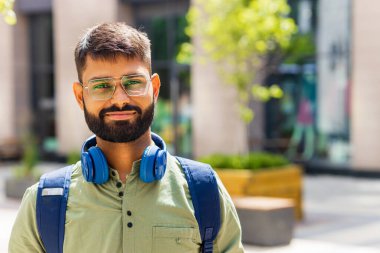 Mavi kulaklıklı ve sırt çantalı Hintli öğrenci güneşli bir güne bakıyor. .