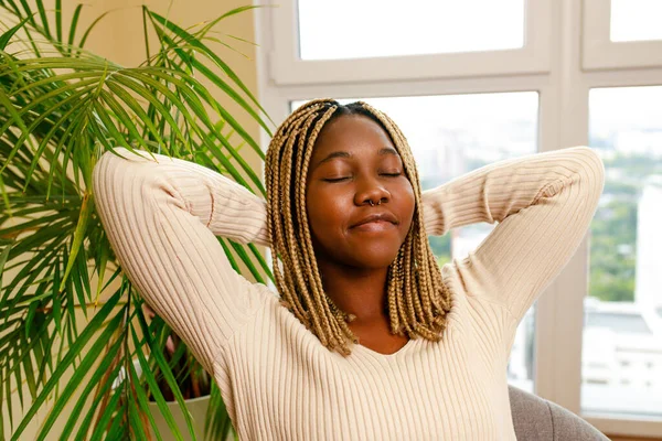 hispanic woman closed eyes chilling on sofa and breathing near green plant at home.