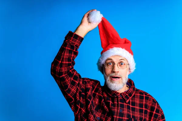 an elderly and well-groomed man in a plaid shirt and santa hat in blue studio background .