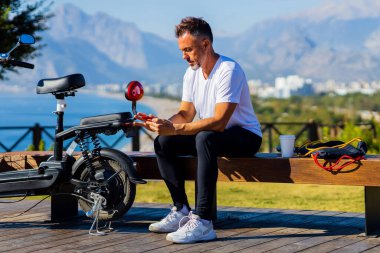 Emekli, mutlu, gri sakallı bir adam, doğa sporundan sonra dinleniyor. Dağ ve sahil manzaralı, eko scooter 'ın yanında..