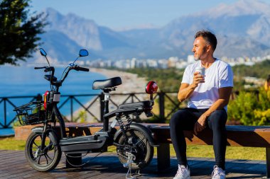Beyaz tişörtlü yakışıklı adam eko scooter 'ın yanında oturuyor. Tepelerde güzel manzarası var ve kahve içiyor..