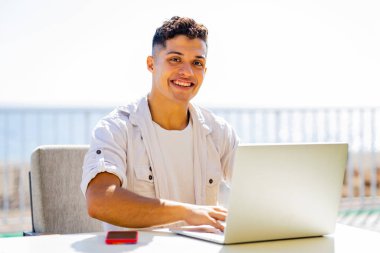 Kumsaldaki kafede dizüstü bilgisayarla çalışan beyaz tişörtlü genç bir adam..