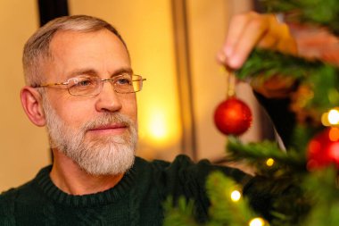 Yeşil örgülü yaşlı bir adam Noel ağacını süslüyor..