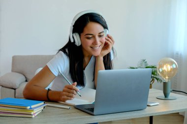Çevrimiçi eğitim kavramı. Genç, güzel esmer bir kadın online bir ders izliyor, defterine bir şeyler yazıyor. Evde dizüstü bilgisayarla çalışan kız öğrencinin portresi. Kapat, kopyala boşluk, arkaplan.