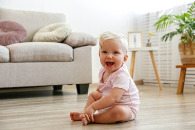 Oturma odasının ahşap zemininde oturan bir yaşındaki bir çocuğun portresi. İç mimari. Evinde pembe sürüngenli sevimli sarışın kız. Kapat, metin için boşluk kopyala.