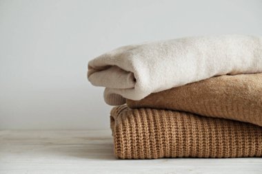 Beyaz ahşap masa üzerine yığılmış farklı örgü desenleri olan bir grup sıcak pastel renkli süveter, beyaz duvar arka planı. Sonbahar mevsimi örgü. Kapat, boşluğu kopyala.