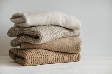 Beyaz ahşap masa üzerine yığılmış farklı örgü desenleri olan bir grup sıcak pastel renkli süveter, beyaz duvar arka planı. Sonbahar mevsimi örgü. Kapat, boşluğu kopyala.