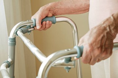 Huzurevinde yaşlı bir kadının kırışık elleriyle yürüyen çerçeveyi tutarken çekilmiş görüntüsü. Kıdemli bayan metal yürüteçlerin saplarını tutuyor. İç arkaplan, kopyalama alanı, kapat.