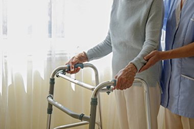 Huzurevinde yaşlı bir kadının kırışık elleriyle yürüyen çerçeveyi tutarken çekilmiş görüntüsü. Kıdemli bayan metal yürüteçlerin saplarını tutuyor. İç arkaplan, kopyalama alanı, kapat.