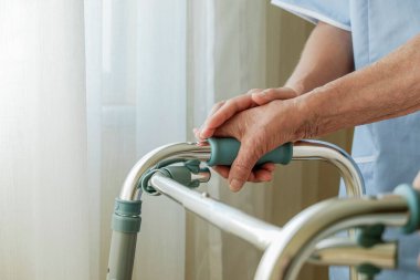 Huzurevinde yaşlı bir kadının kırışık elleriyle yürüyen çerçeveyi tutarken çekilmiş görüntüsü. Kıdemli bayan metal yürüteçlerin saplarını tutuyor. İç arkaplan, kopyalama alanı, kapat.