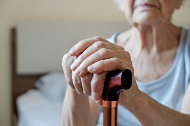 Huzurevinde oturan yaşlı bir kadının eli buruşuk baston tutarken yakın plan fotoğrafı. Metal yardım çubuğu tutan son sınıf öğrencisi bayan. İç arkaplan, kopyalama alanı