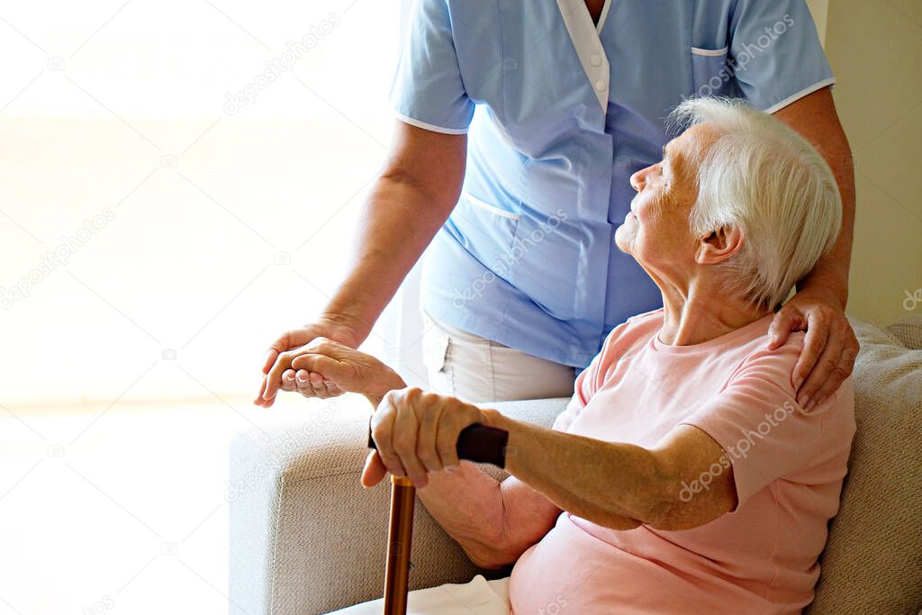 Senior woman holding quad cane handle in elderly care facility. Hospital nurse comforting mature female in nursing home, sitting with walking stick. Background, close up on hands with wrinkled skin.