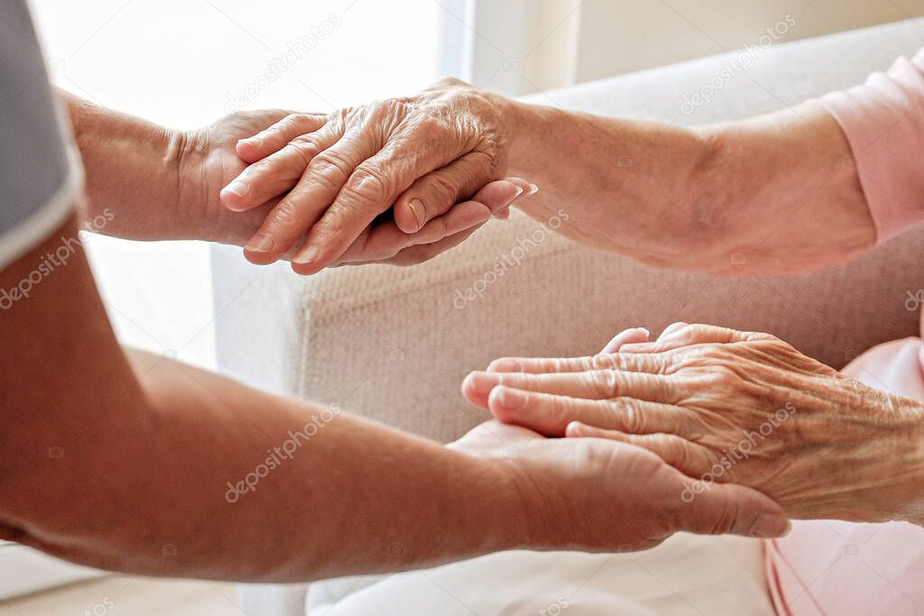 Mature female in elderly care facility gets help from hospital personnel nurse. Senior woman with aged wrinkled skin & care giver, hands close up. Grand mother everyday life. Background, copy space.