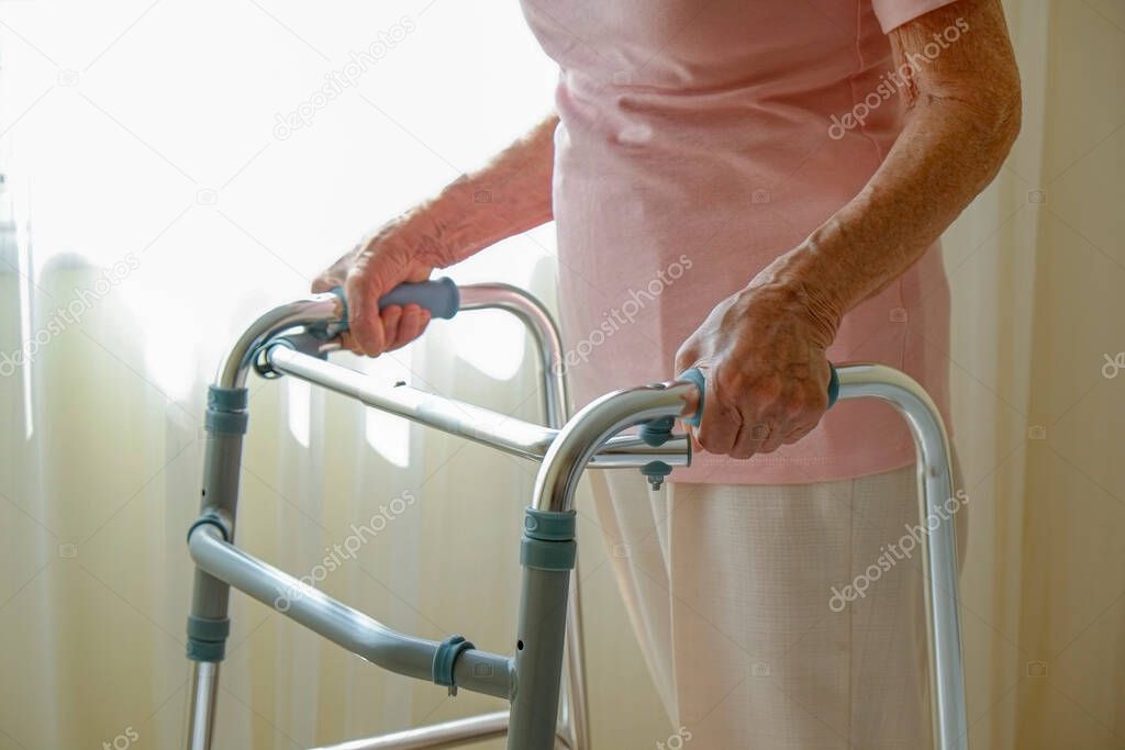 Cropped shot of an elderly woman in nursing home room holding walking frame with wrinkled hand. Senior lady grabbing metal walker's handles. Interior background, copy space, close up.