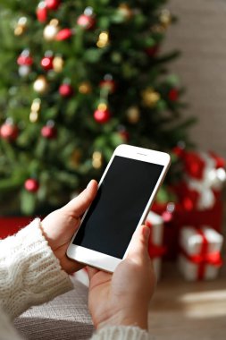Süslü Noel ağacının ve altında hediyelerin olduğu boş ekranlı bir telefonu tutan bir kadın eli. Birinci şahıs görüşü. Kapat, kopyala boşluk, arkaplan.