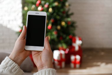 Süslü Noel ağacının ve altında hediyelerin olduğu boş ekranlı bir telefonu tutan bir kadın eli. Birinci şahıs görüşü. Kapat, kopyala boşluk, arkaplan.