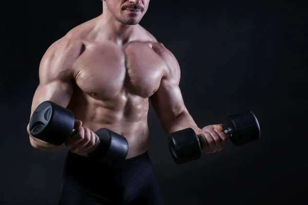 Professional bodybuilder performing biceps curls exercise with dumbbells over isolated black background. Studio shot of a male fitness model pumping iron. Close up, copy space.