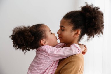 Küçük ve büyük kız kardeş evde birlikte vakit geçiriyorlar. Farklı yaşlarda iki siyah kız kucaklaşıp sevgi gösteriyorlar. Siyahi dişi kardeşler eğleniyor ve kaynaşıyorlar. Arkaplan, kopyala boşluk, kapat.
