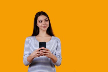 Uzun saçlı, çizgili tişörtlü güzel esmer kadın izole edilmiş sarı arka planda telefonla poz veriyor. Cep telefonunda daktilo eden kadın modelin portresi. Kapat, boşluğu kopyala.