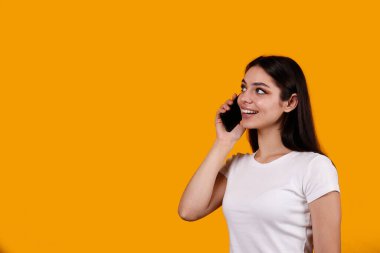 Beyaz tişörtlü, telefonda konuşan ve gülümseyen esmer kadının portresi. Neşeli kadın model, eğlenceli bir telefon görüşmesi yapıyor. İzole edilmiş sarı arkaplan, kopyalama alanı, kapat.