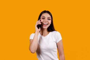 Beyaz tişörtlü, telefonda konuşan ve gülümseyen esmer kadının portresi. Neşeli kadın model, eğlenceli bir telefon görüşmesi yapıyor. İzole edilmiş sarı arkaplan, kopyalama alanı, kapat.